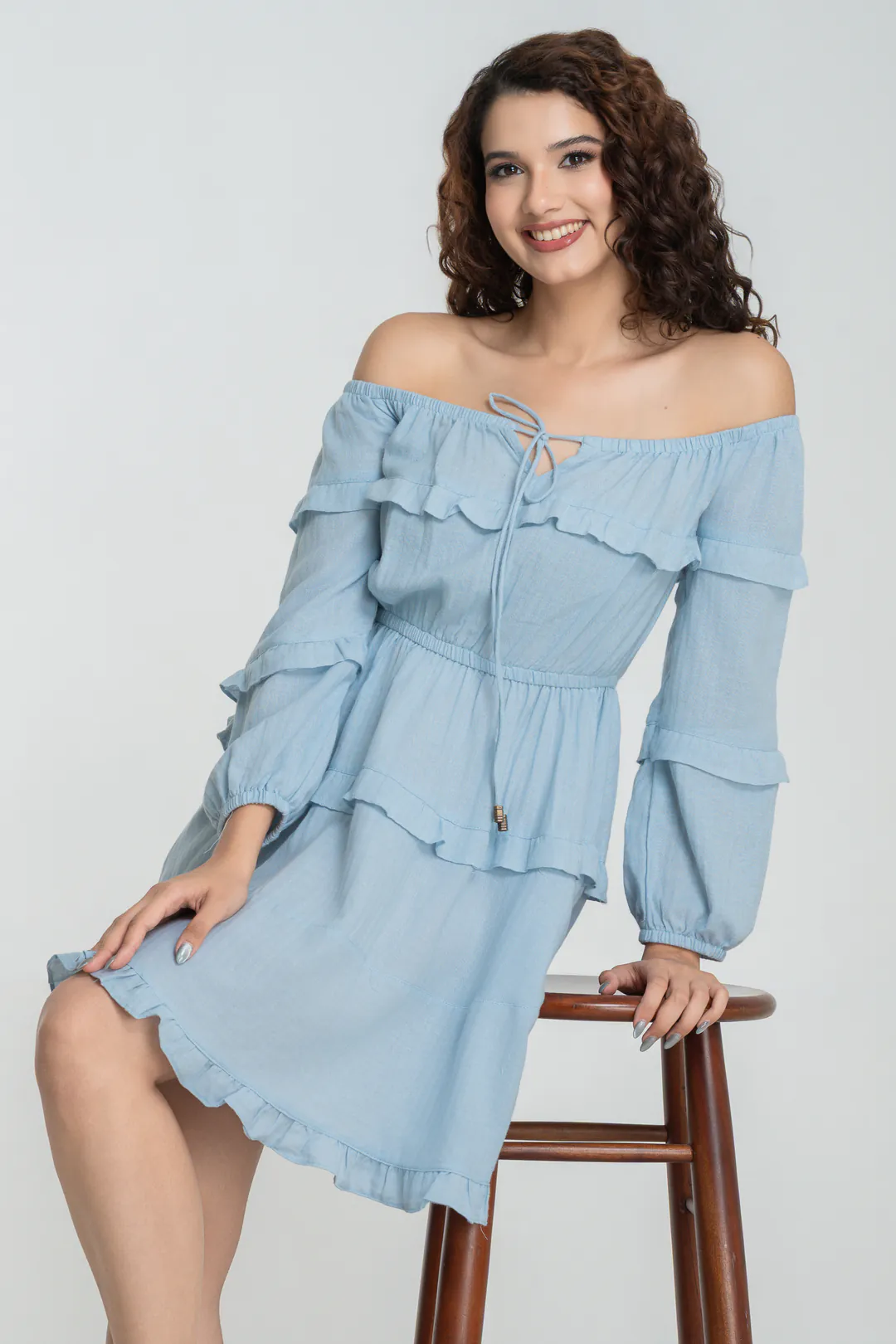 Model seated on a wooden stool in a light blue off-shoulder tiered ruffle dress